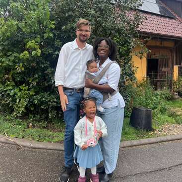 Uma família alegre de quatro pessoas está do lado de fora de uma casa. Os pais sorriem, segurando um bebê, enquanto o filho mais velho, usando uma câmera, ri alegremente.