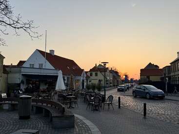 Une scène de rue européenne sereine au coucher du soleil, avec des tables de café en plein air, des routes pavées, de charmants bâtiments, des voitures garées, et une atmosphère tranquille alors que la lumière du jour s'estompe.