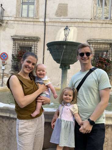 Une famille souriante de quatre personnes pose près d'une fontaine en pierre sur une charmante place. Les parents tiennent leurs jeunes enfants dans leurs bras. Des fleurs et de vieux bâtiments les entourent.