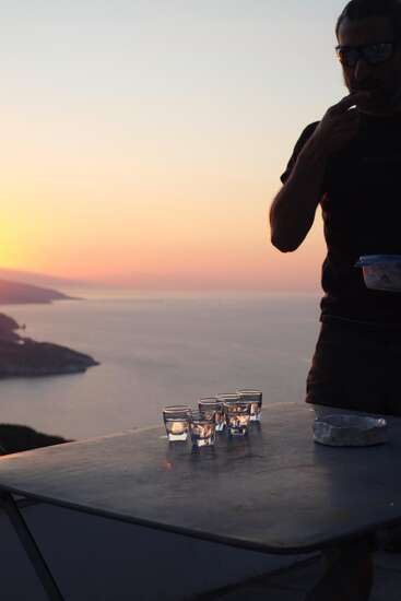 Ein Mann steht bei Sonnenuntergang an einem Tisch und hat einen atemberaubenden Blick aufs Meer. Auf dem Tisch stehen fünf Gläser und ein Teller, die das warme Licht der untergehenden Sonne reflektieren.