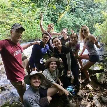 Un groupe de neuf personnes pose joyeusement dans une forêt verdoyante, au bord d'un ruisseau, souriant, faisant des signes de paix et profitant d'une aventure en plein air.