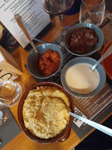 Quatre bols contiennent différents plats italiens, notamment de la polenta crémeuse, une sauce blanche, un ragoût de tomates et un ragoût de bœuf, accompagnés de verres à vin et d'un menu.