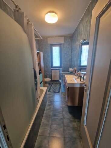 Il s'agit d'une salle de bains étroite, avec du carrelage bleu, une baignoire et un rideau de douche à gauche, un lavabo et un miroir à droite, ainsi que deux fenêtres.