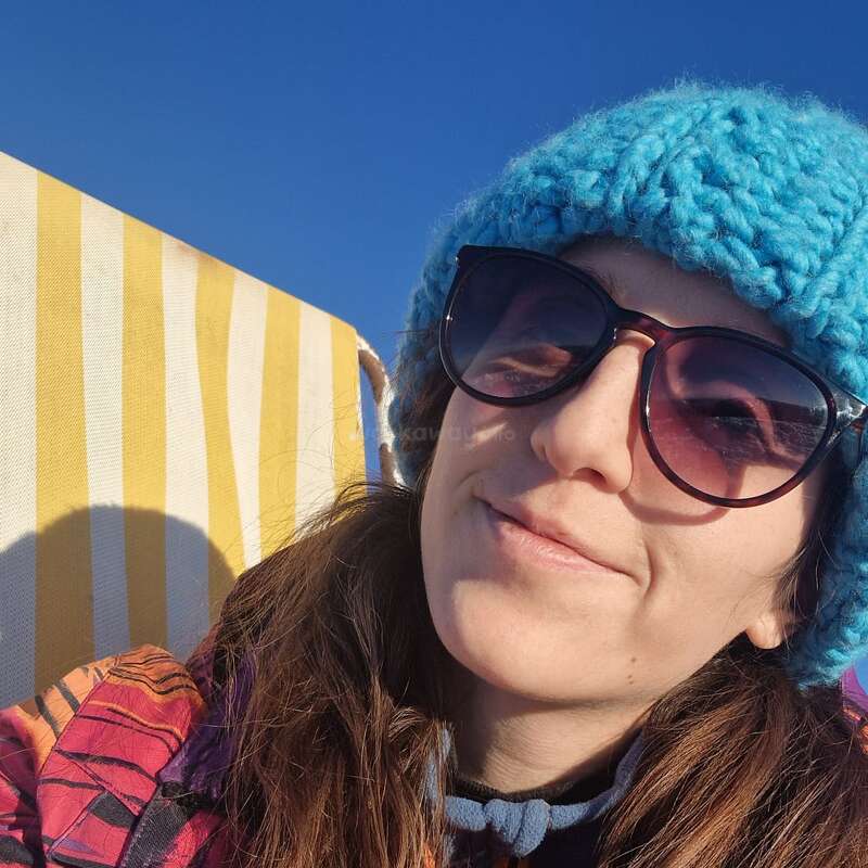 Une femme coiffée d'un bonnet en tricot bleu vif et portant de grandes lunettes de soleil sourit à l'appareil photo. Assise sur une chaise à rayures jaunes, elle profite d'une journée ensoleillée et claire.