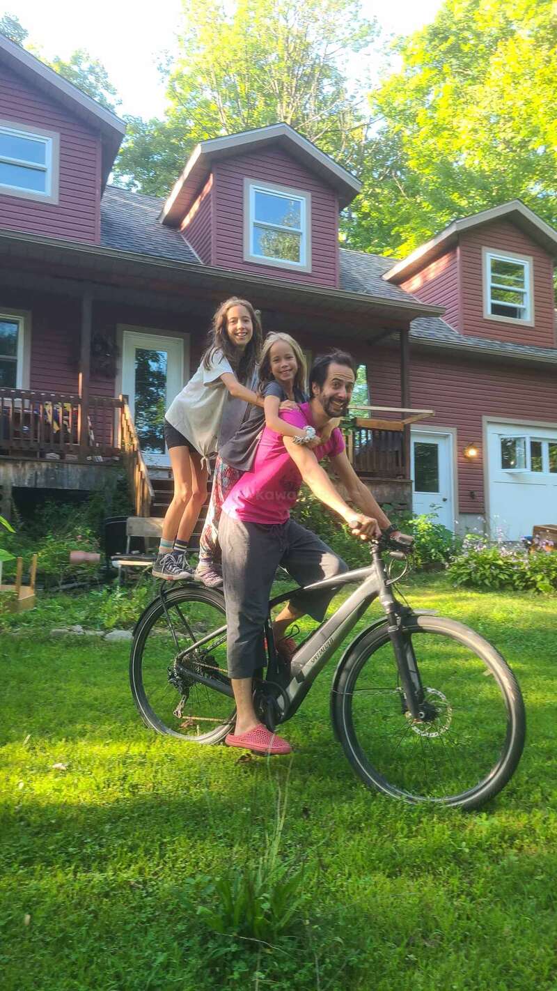 A man rides a bicycle on grass with two smiling children standing behind him. They are in front of a red house surrounded by green trees.