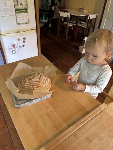 Un niño pequeño, de pie ante una mesa de madera, examina con curiosidad un pan redondo recién horneado que descansa sobre papel pergamino en una cocina cálida y hogareña.