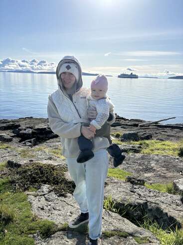 Una mujer, abrigada, sostiene a un bebé con un gorro rosa junto a un pintoresco lago. Luz solar brillante, orilla rocosa, hierba verde y un ferry lejano.