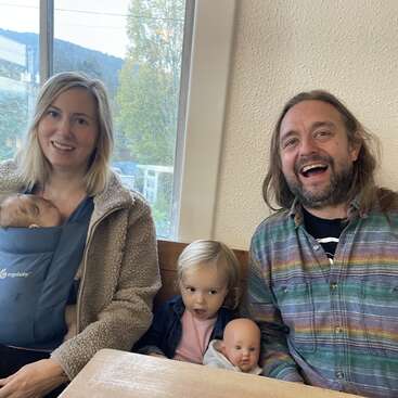 Uma família sorridente está sentada em um ambiente interno. A mãe segura um bebê em um carrinho, o pai está rindo e uma criança segura uma boneca, parecendo surpresa.