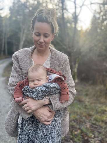 Una mujer de pie al aire libre en un camino, sostiene suavemente a un bebé dormido envuelto en una manta. Los árboles y la suave luz natural crean un ambiente tranquilo.