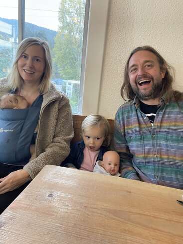 Una familia sonriente se sienta junta en casa. La madre lleva a un bebé en un portabebés, el padre se ríe y un niño pequeño sostiene una muñeca con cara de sorpresa.