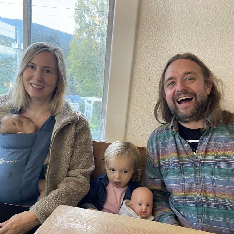 Eine lächelnde Familie sitzt in einem Haus zusammen. Die Mutter hält ein Baby im Arm, der Vater lacht, und ein Kleinkind hält eine Puppe und schaut überrascht.