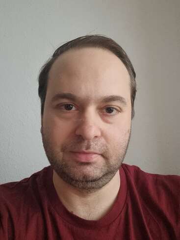 Un homme aux cheveux bruns courts et à la barbe de trois jours porte une chemise bordeaux. Il fait face à la caméra, à l'intérieur, avec une expression neutre et sur un fond uni.