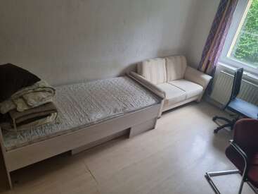 Une chambre simple et bien rangée, avec un lit simple, des draps bien pliés, un canapé beige, deux chaises, une fenêtre avec des rideaux, un parquet clair et des murs blancs.