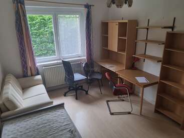 Il s'agit d'une pièce lumineuse dotée de grandes fenêtres, d'un canapé beige, de deux bureaux, de trois chaises, de nombreuses étagères, d'un radiateur, de rideaux et de meubles en bois.