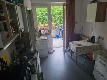 Une cuisine chaleureuse avec une petite table, des étagères ouvertes et un évier. Des portes vitrées donnent sur un balcon agrémenté de plantes, de mobilier d'extérieur et d'un cadre verdoyant.