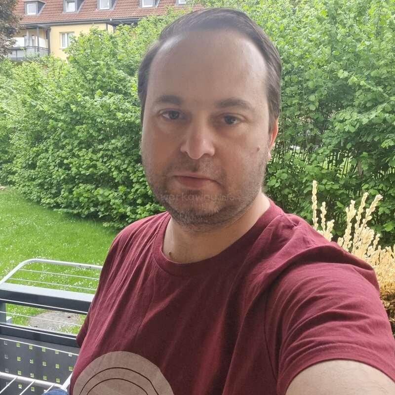 Un homme vêtu d'une chemise bordeaux se tient debout à l'extérieur, près de buissons verts et d'herbe, avec des maisons visibles en arrière-plan. Il regarde l'appareil photo, l'air détendu.