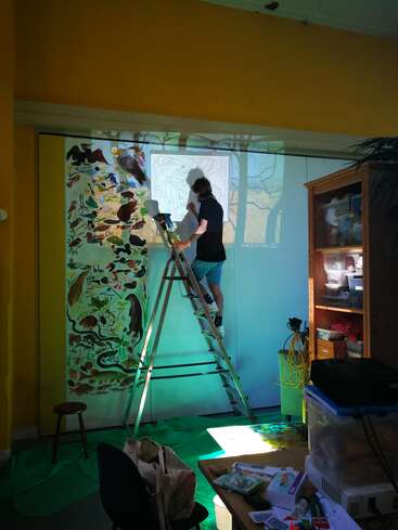 A person stands on a ladder, tracing a projected outline onto a wall for a mural. The room is colorful, filled with art supplies and natural light.