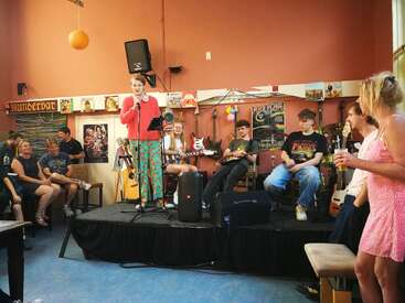 A lively group enjoys a casual music event indoors. People sit and listen as a person in colorful clothes speaks onstage, surrounded by guitars and décor.