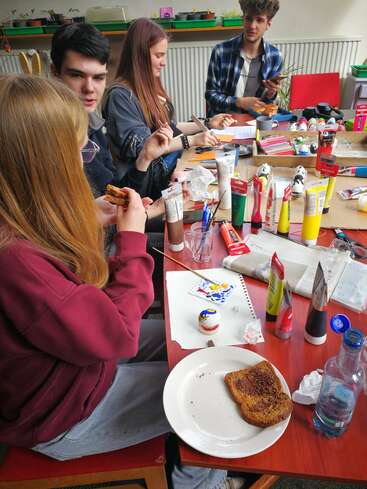 Four young people sit around a table covered in art supplies. They paint eggs, snack on toast, chat, and enjoy a creative, relaxed, and social atmosphere together.