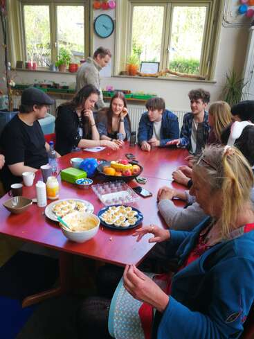 A group of people gathers around a table enjoying food and conversation. Colorful decorations, snacks, and eggs are on the table. The atmosphere appears lively and friendly.