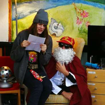 A young person reads from a paper while sitting next to someone dressed as Santa Claus, wearing sunglasses. Colorful artwork and festive objects surround them indoors.