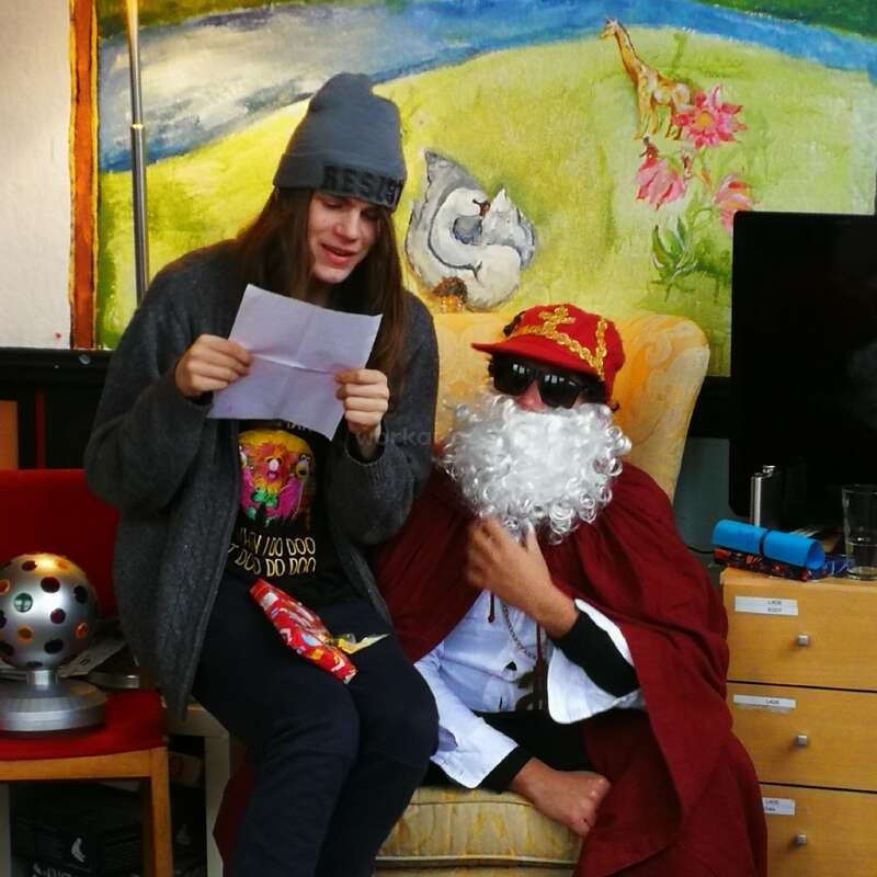 A young person reads from a paper while sitting next to someone dressed as Santa Claus, wearing sunglasses. Colorful artwork and festive objects surround them indoors.