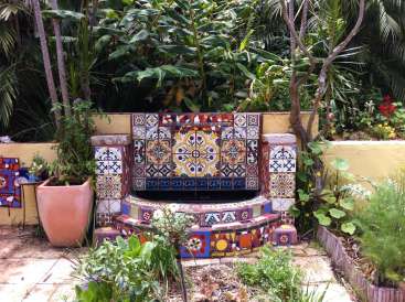 A imagem mostra uma fonte vibrante de azulejos em um jardim exuberante, cercada por plantas e árvores, com uma atmosfera quente e ensolarada.