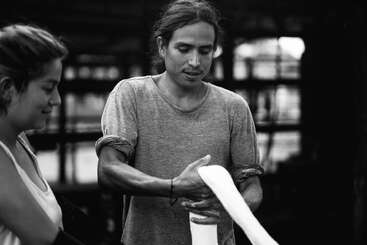 L'image représente un homme aux cheveux longs, vêtu d'un t-shirt gris, tenant un objet blanc, probablement un morceau de papier ou de tissu, devant un arrière-plan flou.
