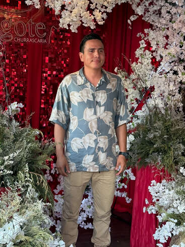 A man stands amidst white flowers and greenery, set against a rich red curtain backdrop. He wears a floral shirt and khaki pants, smiling gently.