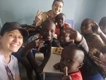 Un grupo de niños sonrientes y dos adultos posan alegremente juntos en un aula. Todos parecen felices, algunos muestran signos de la paz y señalan juguetonamente a la cámara.