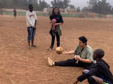 Un grupo de personas se reúne en un polvoriento campo de fútbol. Una persona está sentada, sosteniendo un balón casero, mientras otras permanecen de pie y observan, entablando conversación.