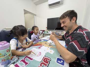Un hombre sonriente se sienta en una mesa con dos niñas pequeñas, dibujando y coloreando juntos. El ambiente es alegre y creativo, con materiales artísticos esparcidos por todas partes.