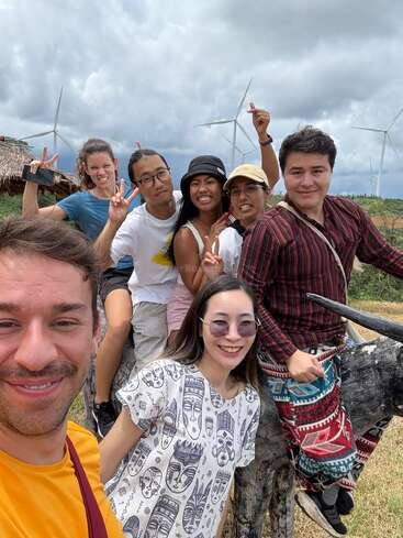 Un grupo de seis amigos sonríe al aire libre, posando con signos de la paz sobre una escultura animal de madera. Las turbinas eólicas y el cielo nublado crean un ambiente animado y aventurero.