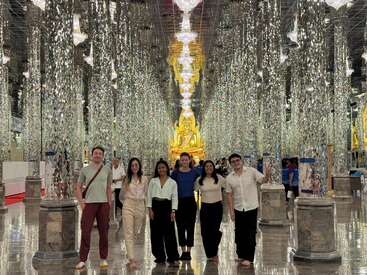 Un grupo de siete personas permanece descalzo en el interior de un deslumbrante templo adornado con pilares reflectantes, lámparas de araña y una gran estatua dorada de Buda al fondo.