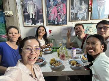 Seis amigos disfrutan alegremente de una comida juntos en un animado restaurante, sonriendo a la cámara. Retratos de miembros de la realeza cuelgan de la pared detrás de ellos.