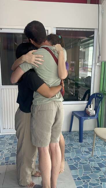 Tres personas están muy juntas, abrazadas. El escenario parece un interior, con suelo de baldosas, sillas de plástico y una caja de pañuelos a la vista.