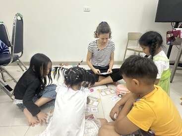 Una mujer y cuatro niños sentados en el suelo en círculo, jugando con fichas y juguetes educativos, parecen disfrutar juntos de una actividad de aprendizaje divertida e interactiva.