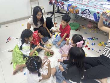 Un grupo de niños pequeños y dos adultos están sentados en el suelo, comiendo bocadillos y jugando con juguetes en un aula alegre y bien iluminada.