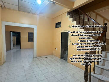 Esta imagen muestra una zona común sencilla, con suelo de baldosas, escaleras, paredes de color beige y una puerta. El texto la describe como un espacio compartido donde viven los voluntarios.