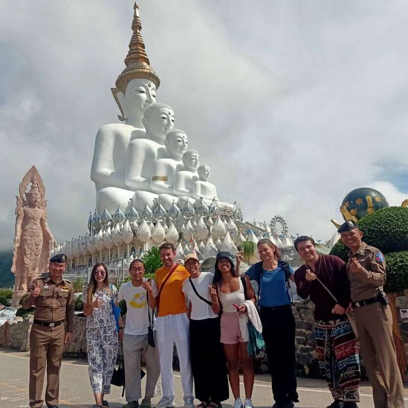 Un grupo de personas sonrientes posa ante las impresionantes cinco estatuas blancas de Buda del templo Wat Pha Sorn Kaew, con exuberante vegetación y cielo nublado.