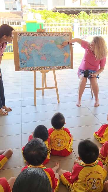 Un enseignant montre une carte du monde accrochée à un chevalet tandis que des enfants vêtus d'uniformes identiques sont assis par terre et écoutent attentivement pendant un cours de géographie en plein air.