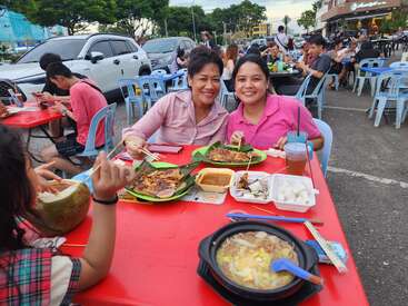 Two women are smiling at an outdoor restaurant, enjoying various local dishes and drinks on a red table. The atmosphere is lively with many diners around.