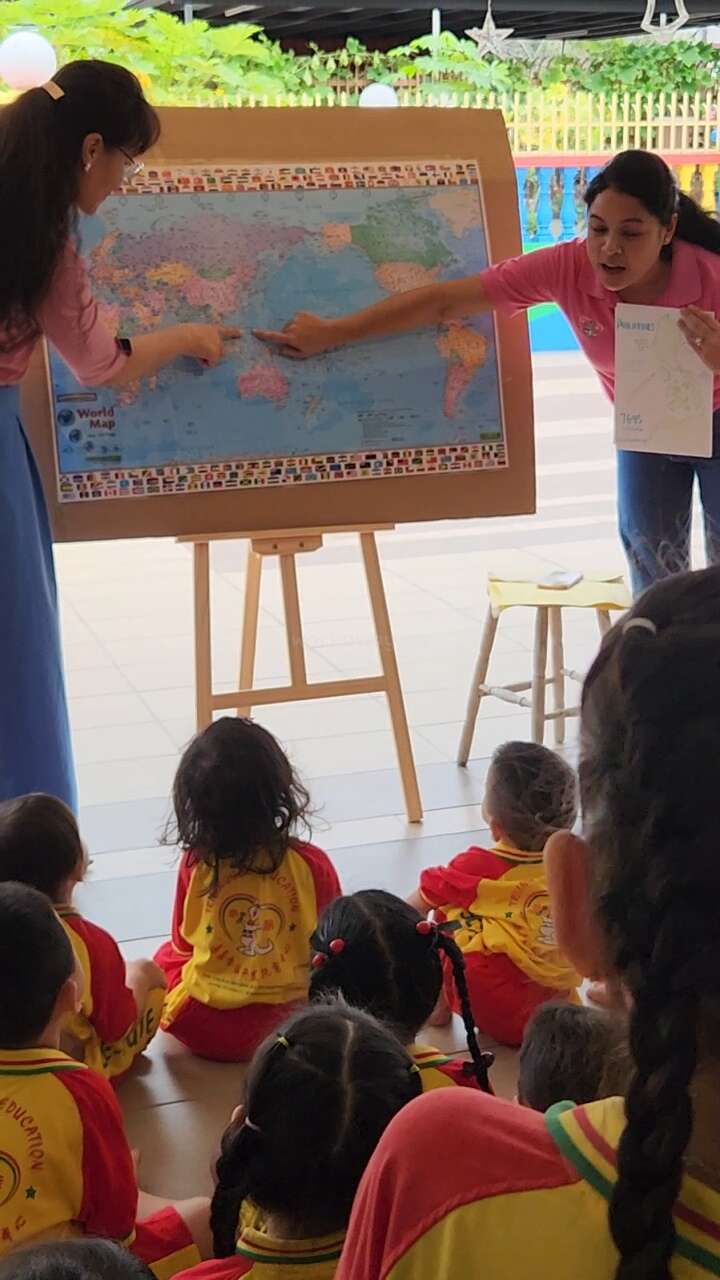 Two teachers point at locations on a world map, engaging a group of young children in yellow uniforms, seated attentively, during an interactive geography lesson.