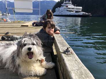 A imagem retrata uma cena serena de uma criança e um cachorro em uma doca, com um barco ao fundo, em um pitoresco cenário montanhoso.