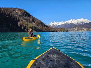 A person kayaks across a stunning turquoise lake, surrounded by forested hills and snow-capped mountains under a clear blue sky, enjoying peaceful nature.