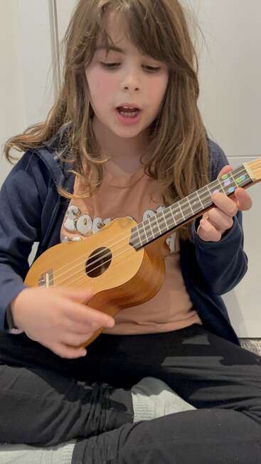 A imagem mostra uma jovem com longos cabelos castanhos, tocando um ukulele sentada no chão, vestindo uma jaqueta azul e calça preta.