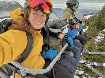 A imagem mostra uma mulher e três crianças em um teleférico, usando equipamentos de inverno, com uma paisagem montanhosa e árvores ao fundo.