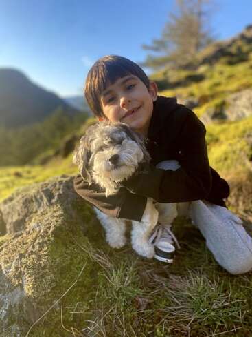 A imagem mostra um menino ajoelhado em uma colina gramada, abraçando um pequeno cachorro branco com manchas escuras, em um sereno cenário montanhoso.