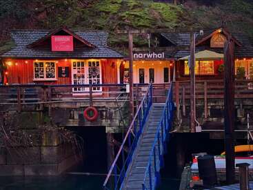 Charming wooden waterfront buildings with glowing lights, a blue ramp leading to the dock, festive windows, lush greenery above, and kayaks below, creating a cozy atmosphere.