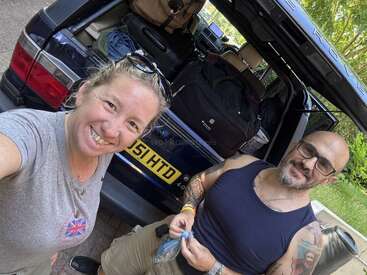 Uma mulher e um homem sorridentes estão ao lado de um porta-malas aberto de um carro cheio de bagagens, prontos para uma viagem de carro, desfrutando juntos de um dia ensolarado ao ar livre.